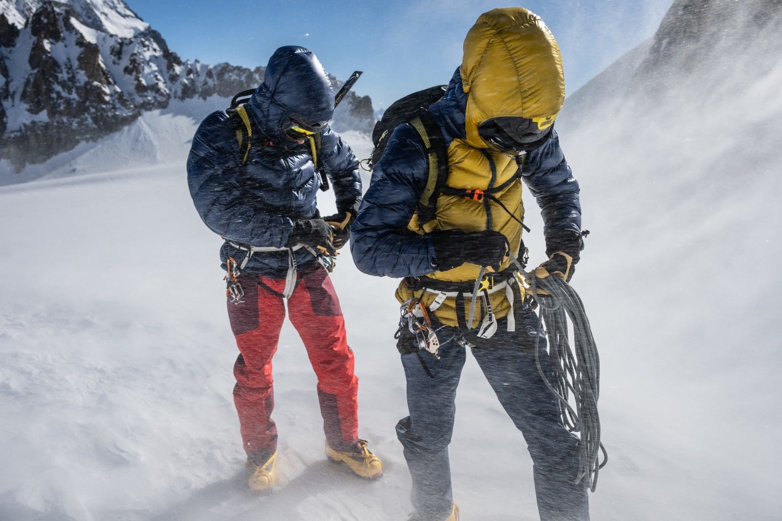 Two roped mountaineers in down jackets and harnesses standing in blowing snow on a glacier